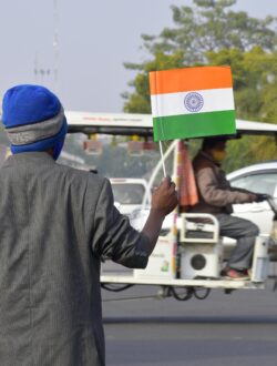 Man holding Indian flag on street corner with moving vehicles.